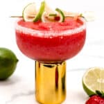 A strawberry margarita with a salted rim, garnished with lime slices, served in a tall gold glass, surrounded by fresh limes and strawberries.