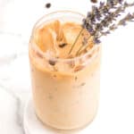 A glass of iced coffee with a sprig of lavender, sitting on a white marble surface, with coffee beans scattered around.