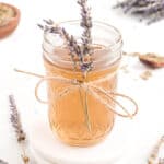 A glass jar filled with amber-colored liquid, decorated with a lavender sprig and twine, on a white coaster surrounded by lavender flowers.