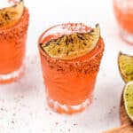 Two glasses of pink cocktails with charred lime garnishes and chili-salted rims, on a white textured surface with scattered spices.