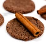 Close-up of chocolate cookies with a cinnamon stick atop one, arranged on a white surface.