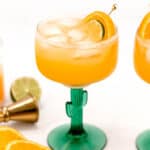 A refreshing orange cocktail served in a green stemmed glass, garnished with an orange slice and presented on a white background with citrus fruits and a cocktail shaker nearby.