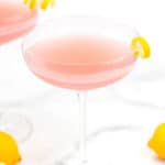 A pink cocktail in a coupe glass garnished with a lemon peel, with a similar drink and whole lemons in the background.