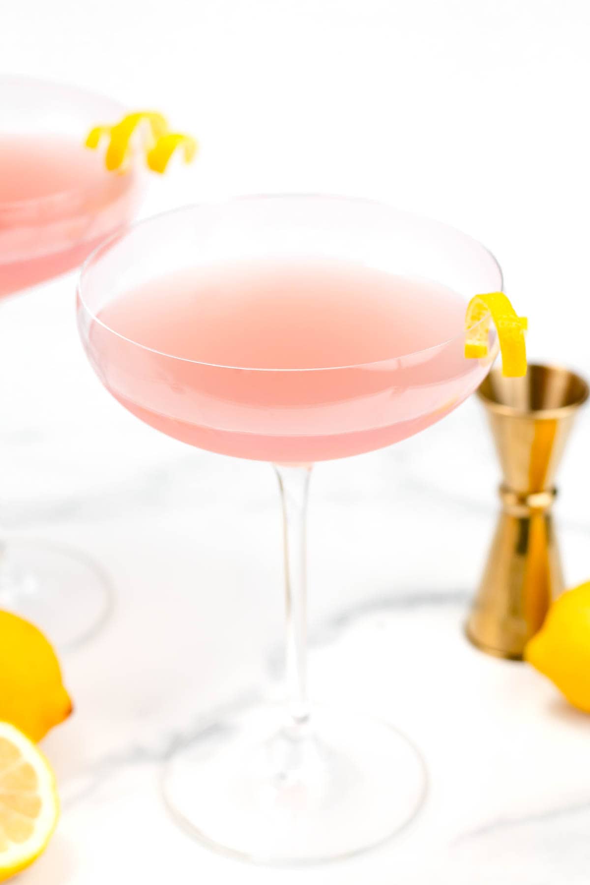 A pink cocktail in a coupe glass garnished with a lemon twist. A jigger and lemons are in the background.
