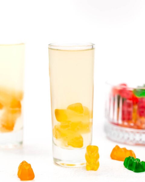 A tall glass of clear liquid with gummy bears at the bottom, surrounded by loose gummy bears and another glass in the background on a white surface.