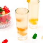 Two glasses of clear liquid with gummy bears at the bottom, on a white surface with a blurred bowl of salad in the background.