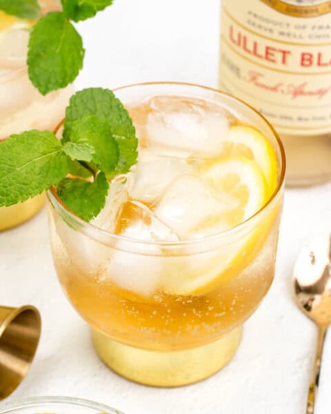 A refreshing cocktail with ice and mint garnish, served in a glass beside a bottle of Lillet Blanc on a white surface.