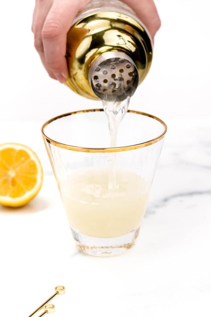 A person pouring a lemon cocktail from a gold shaker into a clear glass on a white surface, with half a lemon and a cocktail pin nearby.