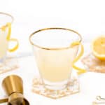A glass of lemonade with a lemon peel garnish, beside a jigger and cocktail stirrer, on a white surface with a subtle marble pattern.