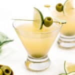 Two dirty martinis in classic glasses garnished with olives and lime wedges, served on a white surface with extra olives and lime slices nearby.