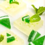 Close-up of square jelly desserts topped with green and white striped candy pieces and fresh mint leaves, arranged on a white surface.