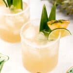 Two pineapple margarita cocktails with ice, garnished with pineapple slices and green leaves, presented on a light background with a pineapple and lime.