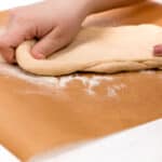 A close-up of a hand kneading dough on a flour-dusted parchment paper.