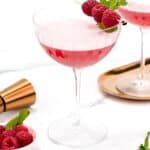 Two raspberry cocktails in coupe glasses garnished with fresh raspberries, accompanied by a shaker and a plate with mint leaves on a white surface.