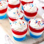 Small cups filled with layered red, white, and blue gelatin dessert topped with whipped cream and red, white, and blue sprinkles. The cups are arranged on a wooden board.
