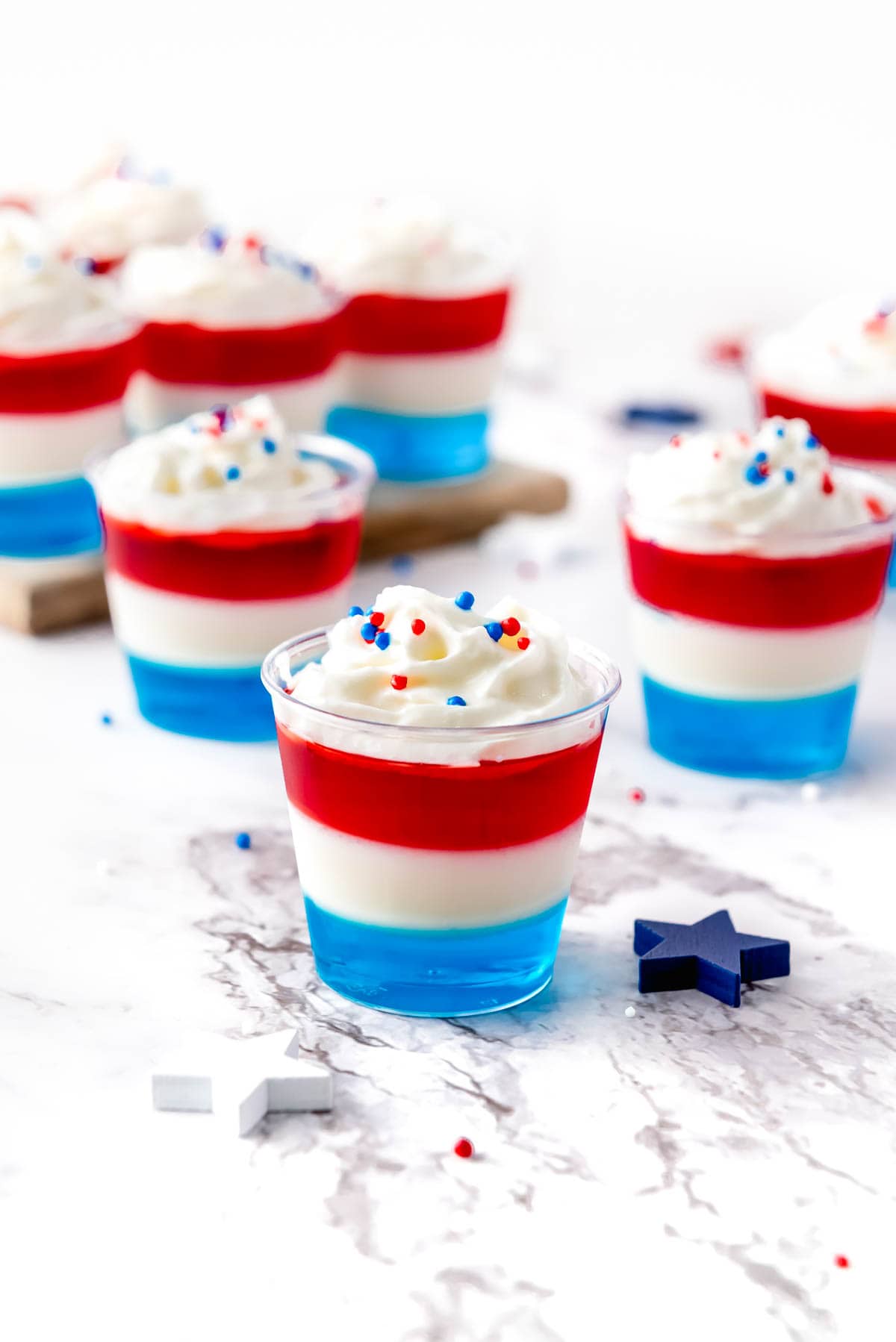 Several small dessert cups with layered red, white, and blue gelatin topped with whipped cream and sprinkles are arranged on a marble surface. Blue star decorations are scattered around them.