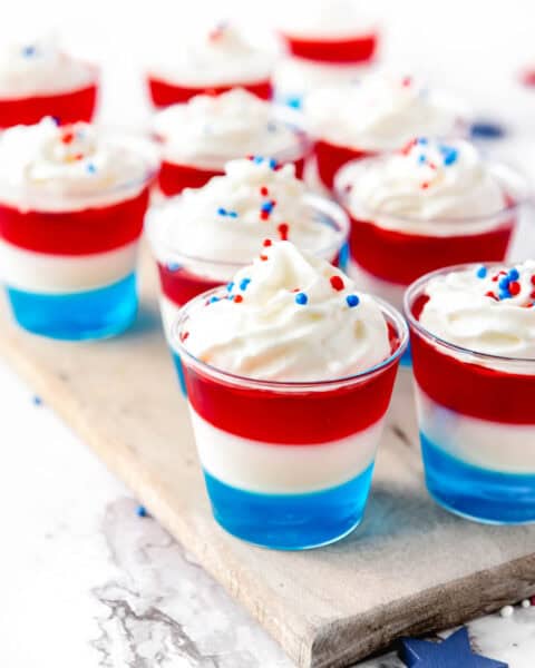 Nine small dessert cups displayed on a wooden board. Each cup consists of three layers colored blue, white, and red, topped with whipped cream and red, white, and blue sprinkles.