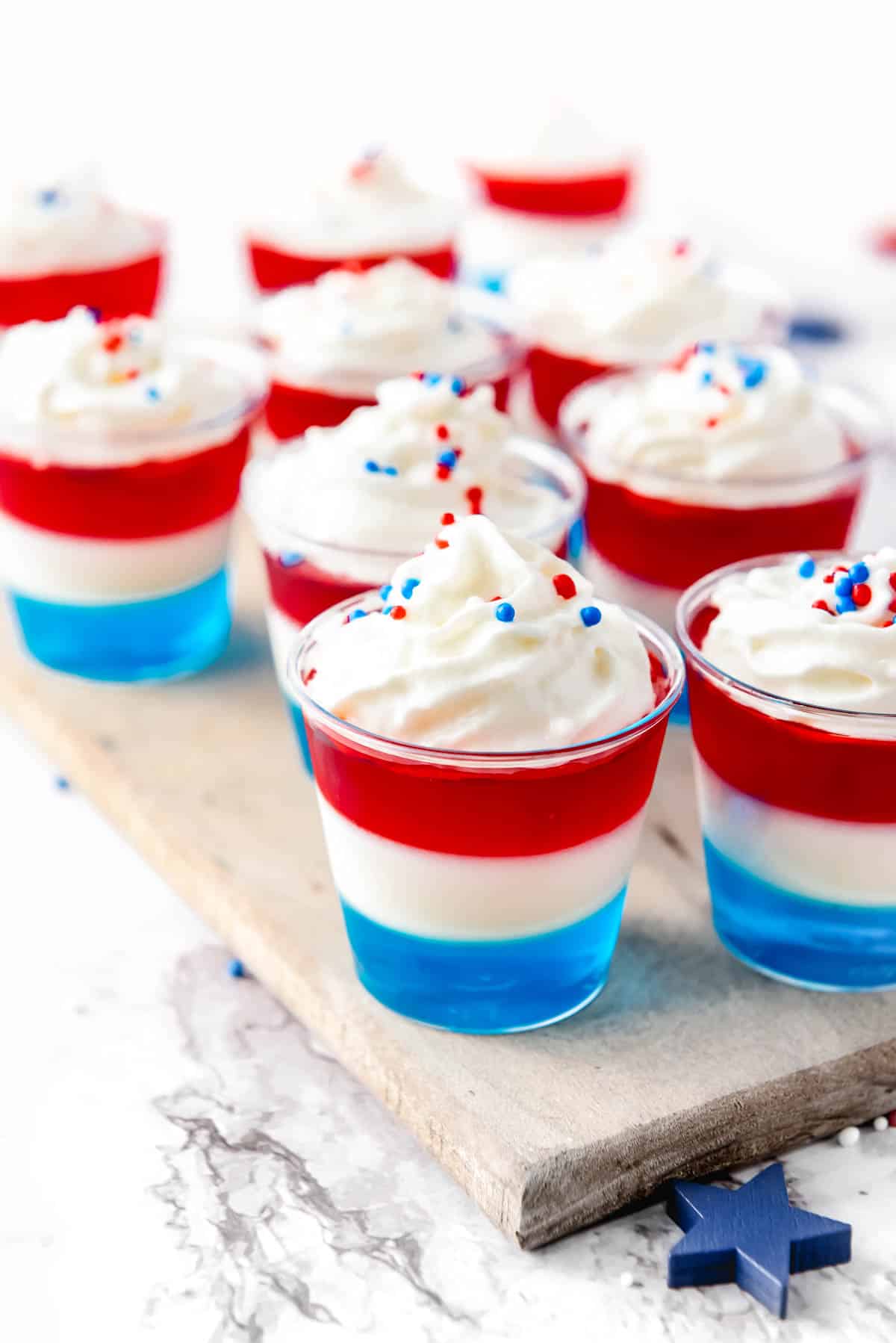 Nine small dessert cups displayed on a wooden board. Each cup consists of three layers colored blue, white, and red, topped with whipped cream and red, white, and blue sprinkles.
