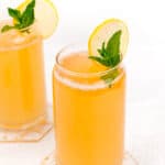Two glass jars of orange-colored drink garnished with lemon slices and mint leaves, placed on hexagonal coasters.