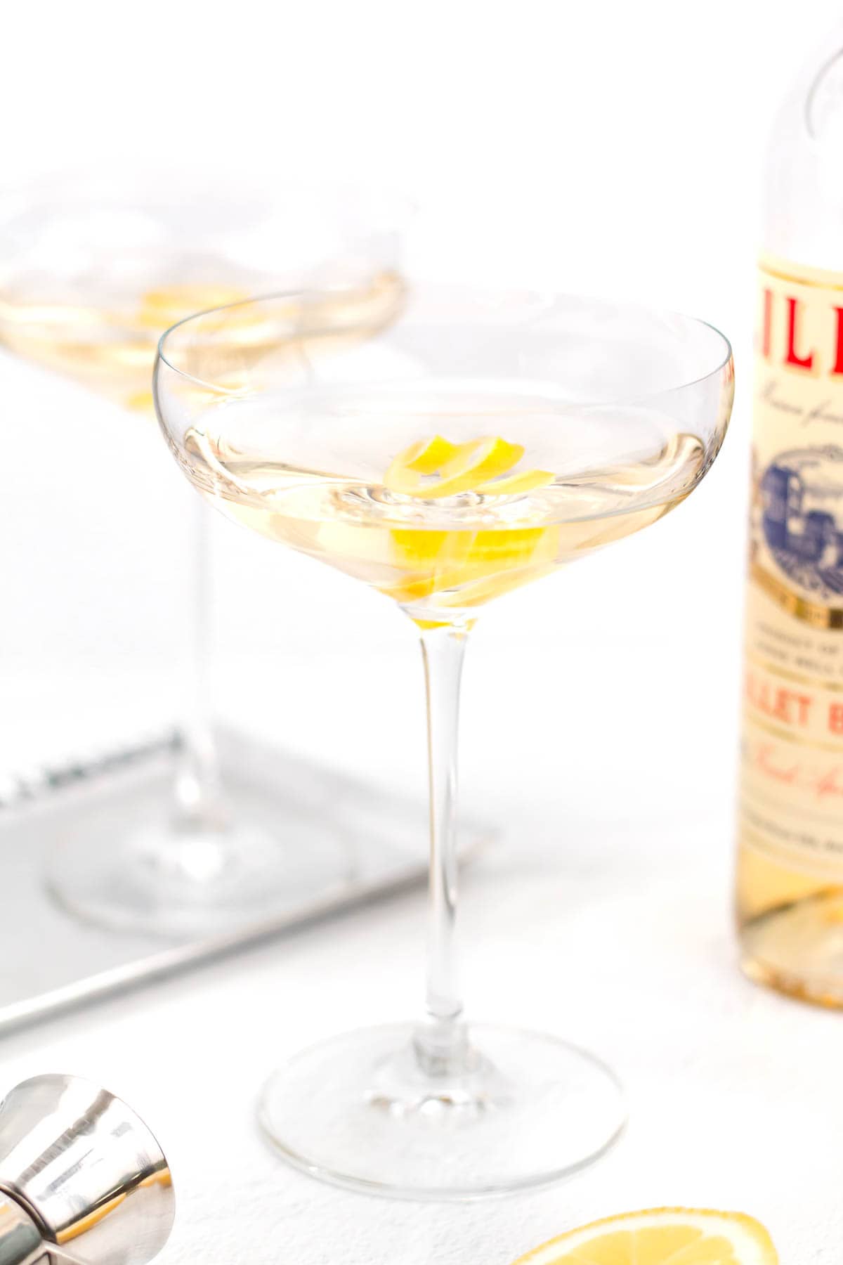 A cocktail in a clear glass garnished with a twist of lemon peel, with another similar glass, a bottle, and lemon slice in the background.