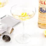 A cocktail glass with a lemon twist garnish, a bottle of Lillet Blanc, a jigger, a slice of lemon, and a serving tray are on a white surface.