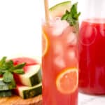 A tall glass of watermelon lemonade with ice, garnished with a lemon slice, mint leaves, and a watermelon wedge. A pitcher of the drink and a cutting board with watermelon pieces are in the background.