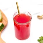 A glass of red watermelon juice with a gold spoon inside, placed next to watermelon slices, a jigger, and green mint leaves on a white surface.