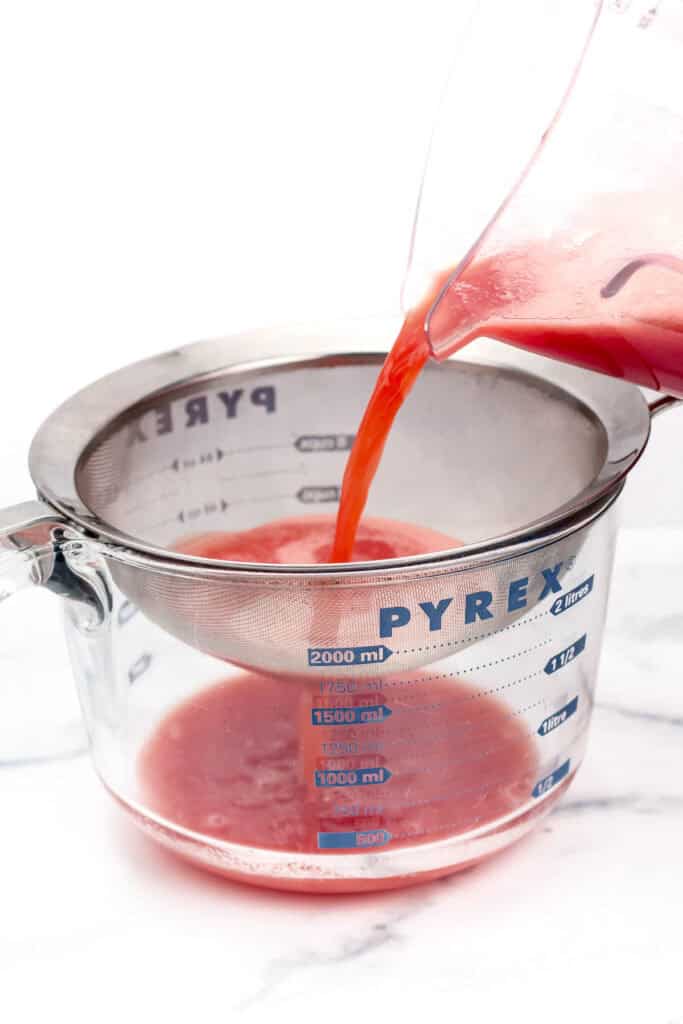 Red liquid being poured through a fine-mesh strainer into a glass measuring cup marked "Pyrex.