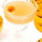A close-up of a cocktail in a decorative glass garnished with a slice of caramelized banana. A gold spoon and a yellow cloth are visible in the background.