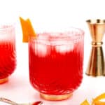 Two red cocktails in ornate glasses garnished with orange peels sit on a white surface. A metal jigger and a spoon with orange peels are beside the drinks.