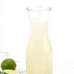 A glass carafe filled with a light yellow liquid, surrounded by whole and halved limes on a white background.