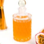 A glass bottle filled with orange liquid sits on a white surface, accompanied by a gold measuring jigger, a sliced passionfruit, and a gold spoon.