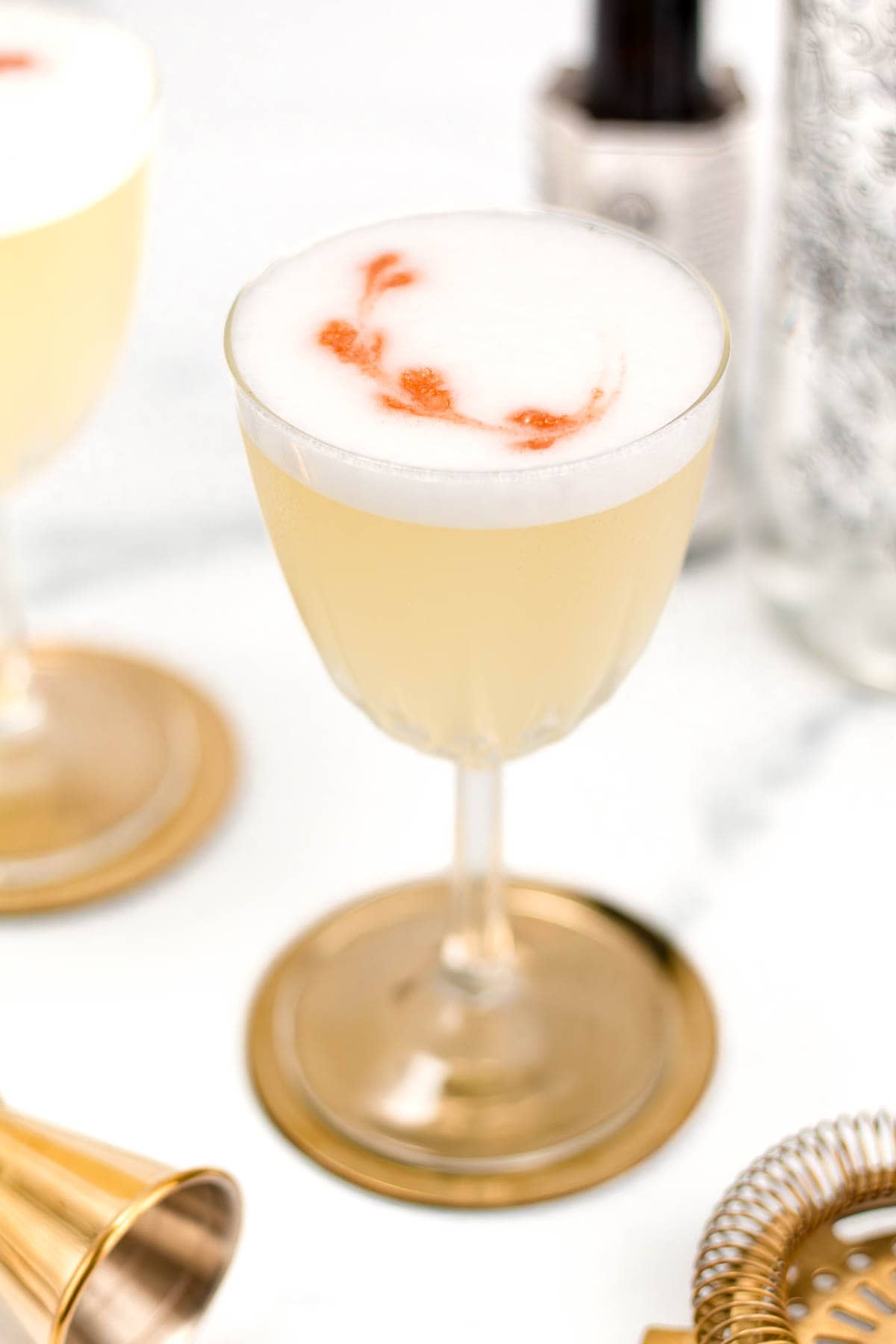 A yellow cocktail with a frothy white top and red swirls, served in a clear glass with a gold base.