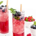 Two tall glasses with red berry drinks, topped with ice, fresh mint, and berries, with bamboo straws. A bowl of assorted berries is in the background.