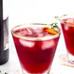 A glass of red iced beverage garnished with an orange slice and a sprig of herb, placed on a white surface with another similar drink in the background.