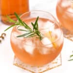 A round glass filled with an iced pink drink garnished with a sprig of rosemary and a lemon twist sits on a coaster, with another identical drink and a pink bottle in the background.