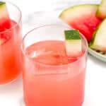 Two glasses of pink-colored watermelon beverages with ice cubes and garnished with watermelon slices. A plate of watermelon slices is visible in the background.