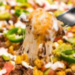 A close-up of a loaded nacho dish with melted cheese, ground beef, corn, jalapeños, diced tomatoes, sour cream, and cilantro. A spoon is lifting a cheesy portion from the dish.