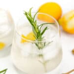 A close-up of a glass of cocktail with ice, garnished with a sprig of rosemary and lemon peel, with a corkscrew and halved lemons in the background.