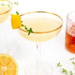 A cocktail garnished with lemon peel and a sprig of thyme in a glass with a gold rim. Additional garnishes and a glass with a reddish liquid are in the background.
