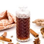A clear, patterned glass bottle filled with dark liquid is surrounded by cinnamon sticks, star anise, cardamom pods, and a piece of ginger root on a white surface.