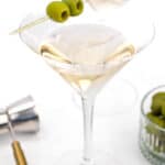 A martini glass filled with a clear beverage, garnished with three green olives on a toothpick. A metal jigger and a small bowl of green olives are seen in the background.