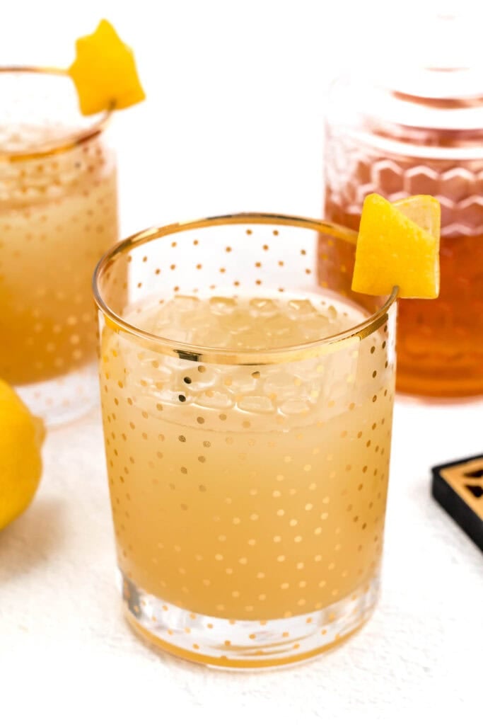 Two glasses of an iced citrus drink with decorative gold dots, garnished with lemon peels. One glass is in the foreground, while the other is partially visible in the background next to a jar and a lemon.