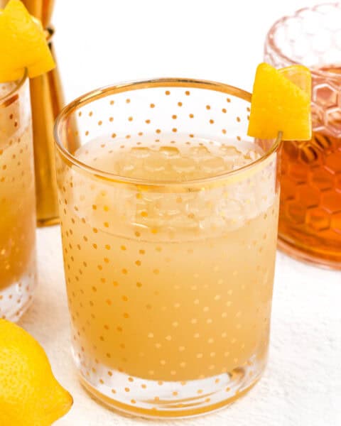 A glass with a yellow, polka-dotted pattern is filled with a light orange drink and ice cubes. A lemon peel garnish is placed on the rim. Lemons and another glass are visible in the background.