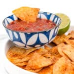 A bowl of salsa with a tortilla chip dipped in it, beside a plate of seasoned tortilla chips and a halved lime.