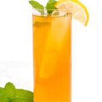 A tall glass of iced tea with a slice of lemon on the rim and mint leaves garnish, placed on a white surface.