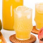 A glass of iced mango lemonade with a lemon slice, set on a round wicker coaster, sits beside a bottle of the same drink, with a mango and lemon nearby on a wooden cutting board.