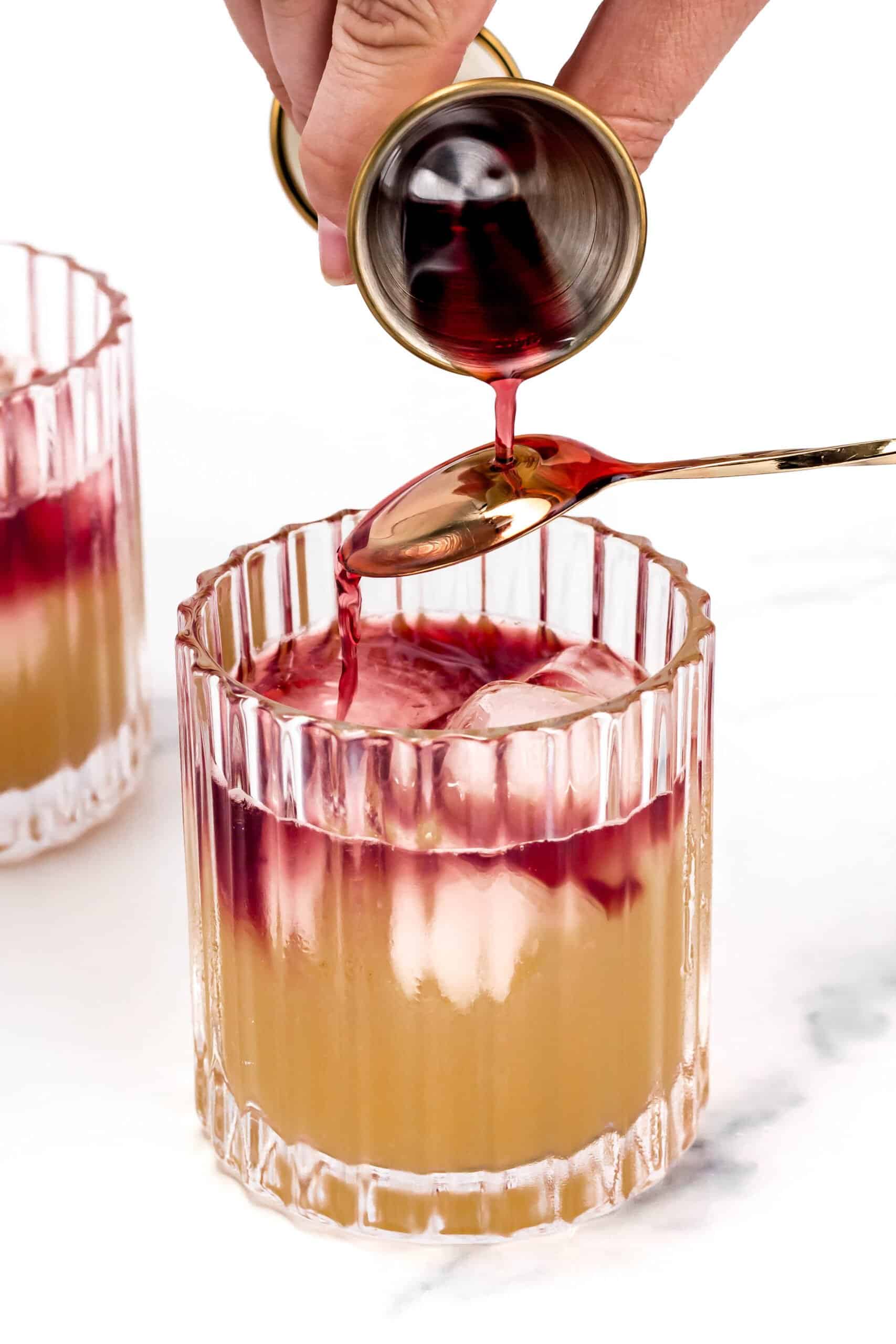 A glass filled with a light-colored drink and ice cubes is being topped with a red liquid being poured from a small gold cup onto a spoon. Another similar glass is in the background.