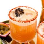 A glass with an orange cocktail garnished with a dried citrus slice and a salted rim, placed on a golden tray with halved passion fruits and a lime nearby.