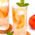 Two tall glasses of iced peach lemonade with lemon slices, peach slices, and mint leaves on top, placed on woven coasters. A fresh peach is visible in the background.
